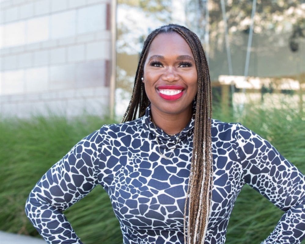 A woman of color smiles and poses for a photo outside of a building.