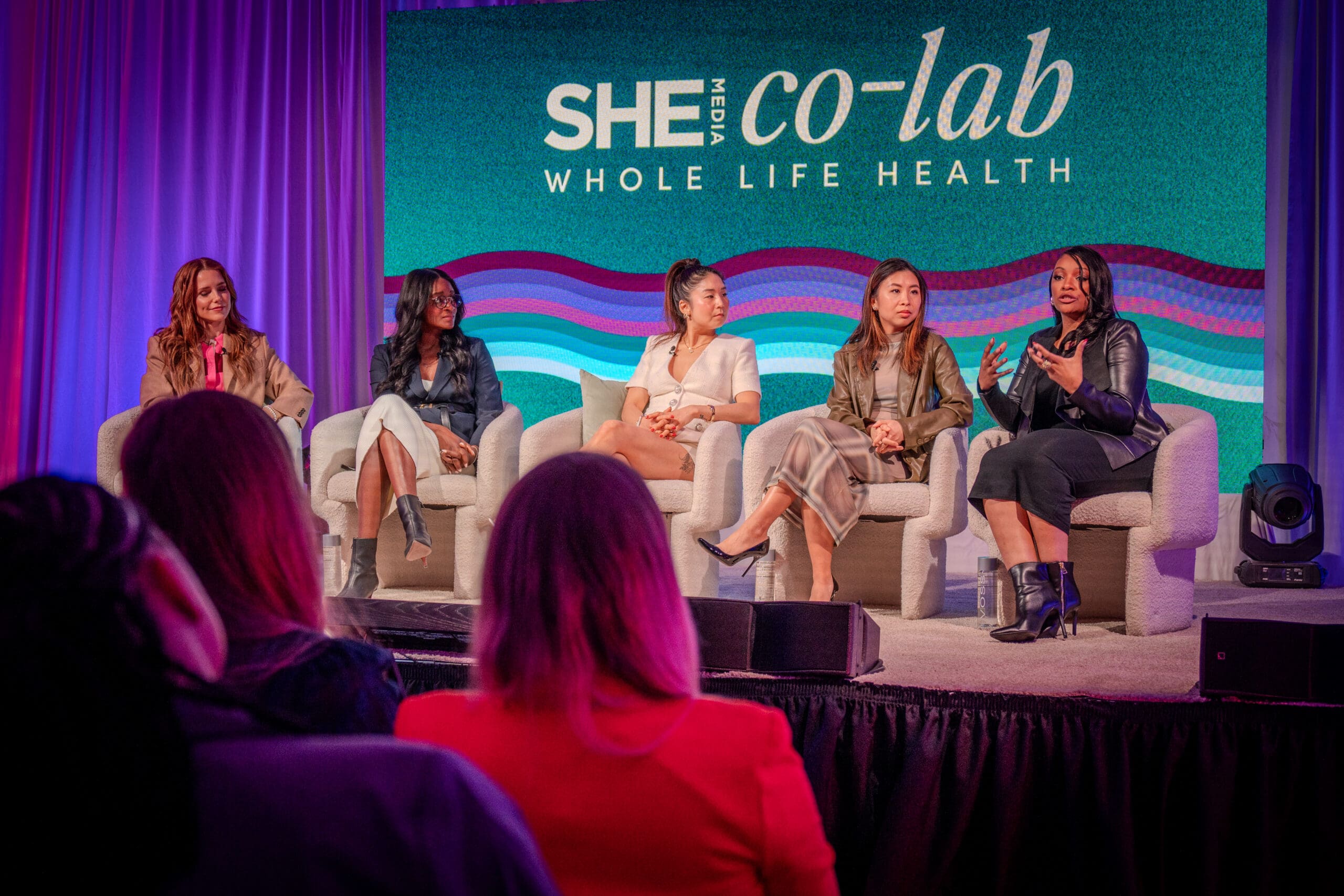Five people sit on couches on a stage speaking to an audience. Behind them is signage for the summit, SHE Media Co-Lab Whole Life Health