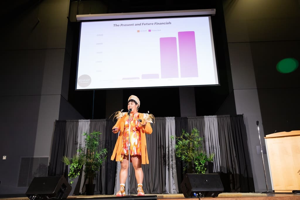 A person stands on an auditorium stage, speaking into a microphone attached to a mic stand. Behind this person is a projector screen displaying a bar chart