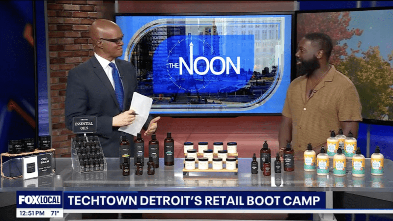 A screenshot of a YouTube clip featuring Scott Boateng talking to Lee Thomas of Fox 2 Detroit. On the news table in front of them are display of Preva Body products. Behind them is a screen that shows a slide for Fox 2 segment "The Noon"
